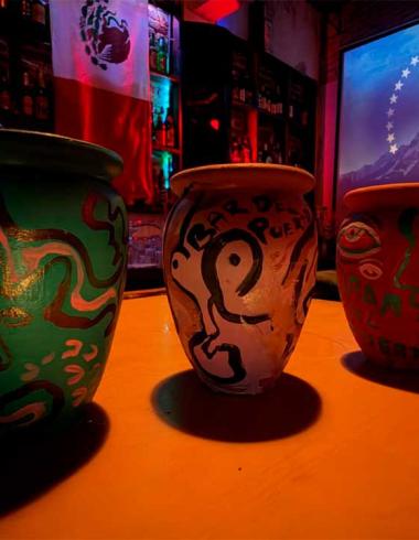 Tres cantaritos de barro pintados a mano sobre la barra del Bar del Puerto, iluminados con luces de colores y con una proyección al fondo.