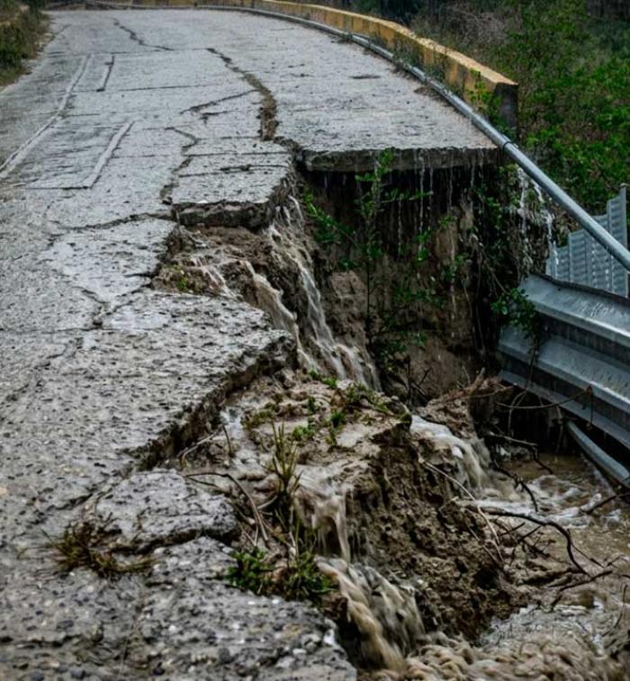 Fotografía de una carretera de concreto rural gravemente colapsada en un acantilado bajo una fuerte lluvia. Una parte significativa del pavimento se ha desmoronado. El agua de lluvia fluye activamente por las grietas y las caras expuestas de la tierra y la roca, y cae sobre un culvert de metal oxidado al pie del colapso. Árboles verdes se alinean en la colina bajo el clima lluvioso.