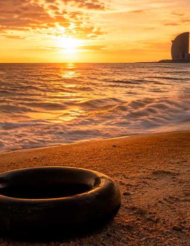Una llanta de cámara de camión descansa sobre la arena de una playa al atardecer. El sol ilumina el horizonte con tonos dorados mientras el mar se extiende hacia una ciudad costera al fondo, evocando un paisaje de memoria, viaje y contemplación.