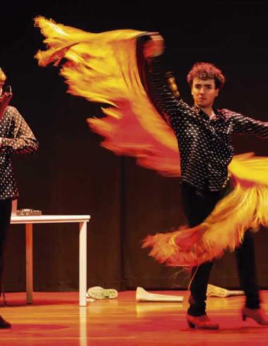Dos artistas se presentan sobre un escenario; uno interpreta un instrumento de viento de madera mientras el otro realiza un movimiento de danza con una larga pieza de tela amarilla y naranja.