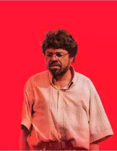 Un hombre de lentes y cabello alborotado con camisa de manga corta mirando de frente sobre un fondo rojo intenso, fotograma de la película Un Poeta de Simón Mesa Soto.