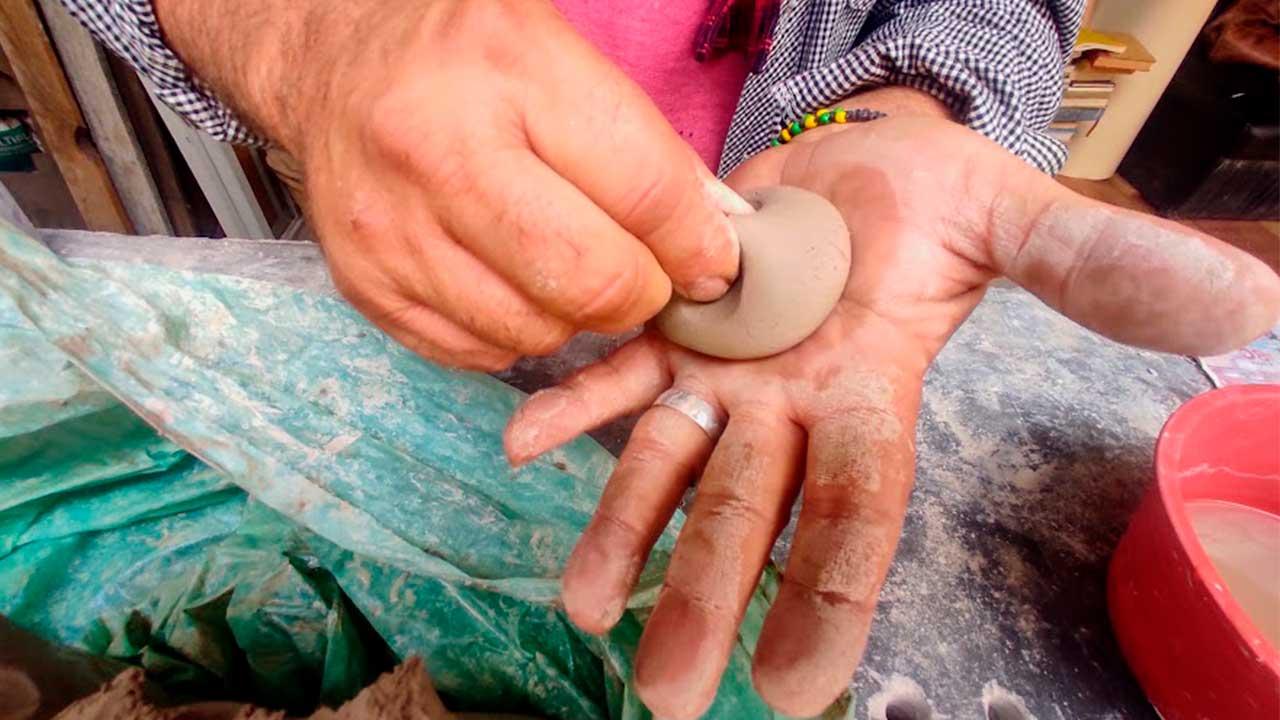 Plano detalle de las manos de un artesano manchadas de tierra mientras moldea una pequeña pieza circular de barro gris en la palma de su mano. Al lado sobre la mesa de trabajo hay un recipiente con agua y plásticos de protección.