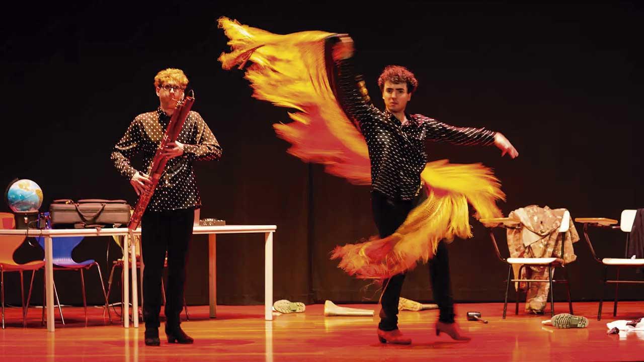 Dos artistas se presentan sobre un escenario; uno interpreta un instrumento de viento de madera mientras el otro realiza un movimiento de danza con una larga pieza de tela amarilla y naranja.