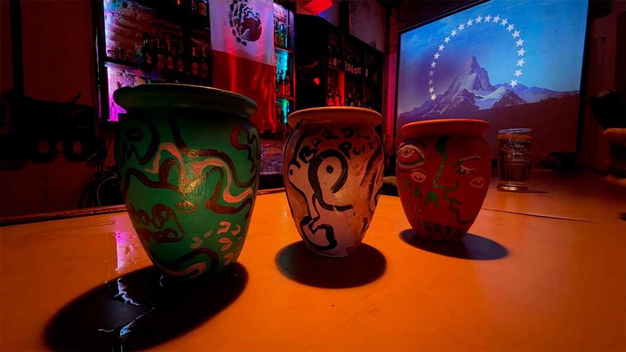 Tres cantaritos de barro pintados a mano sobre la barra del Bar del Puerto, iluminados con luces de colores y con una proyección al fondo.