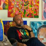 Antonio Tabáres Gallardo sentado en su taller entre pinturas abstractas coloridas, con pinceles y botellas de pintura a su lado, mirando pensativo hacia un lienzo.