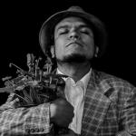 Retrato en blanco y negro del actor y director teatral Pedro Acevedo, con sombrero y saco a cuadros, sosteniendo un ramo de plantas entre sus brazos mientras mantiene los ojos cerrados en una pose introspectiva sobre un fondo negro.