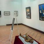 Vista panorámica de la exposición "Orgullo de mis raíces: Copanatoyac" en una sala del Museo Universitario José Juárez. Las pinturas de Miguel Altamirano Velázquez se exhiben en los muros junto con cédulas informativas, mostrando escenas inspiradas en la danza del Tekuanimej y las tradiciones de Copanatoyac, Guerrero.