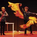 Dos artistas se presentan sobre un escenario; uno interpreta un instrumento de viento de madera mientras el otro realiza un movimiento de danza con una larga pieza de tela amarilla y naranja.