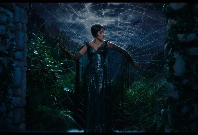 Jennifer López como La Mujer Araña en un escenario teatral, de pie frente a una telaraña gigante que refleja su silueta, vestida con un traje brillante de lentejuelas.