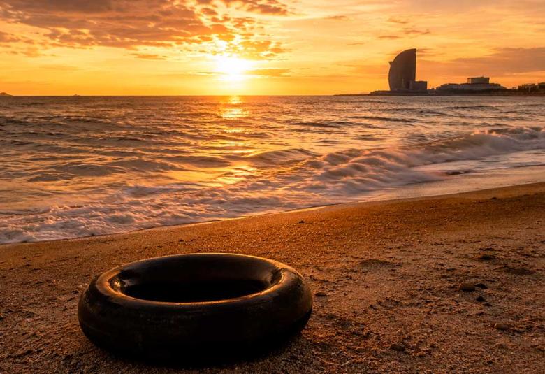 Una llanta de cámara de camión descansa sobre la arena de una playa al atardecer. El sol ilumina el horizonte con tonos dorados mientras el mar se extiende hacia una ciudad costera al fondo, evocando un paisaje de memoria, viaje y contemplación.