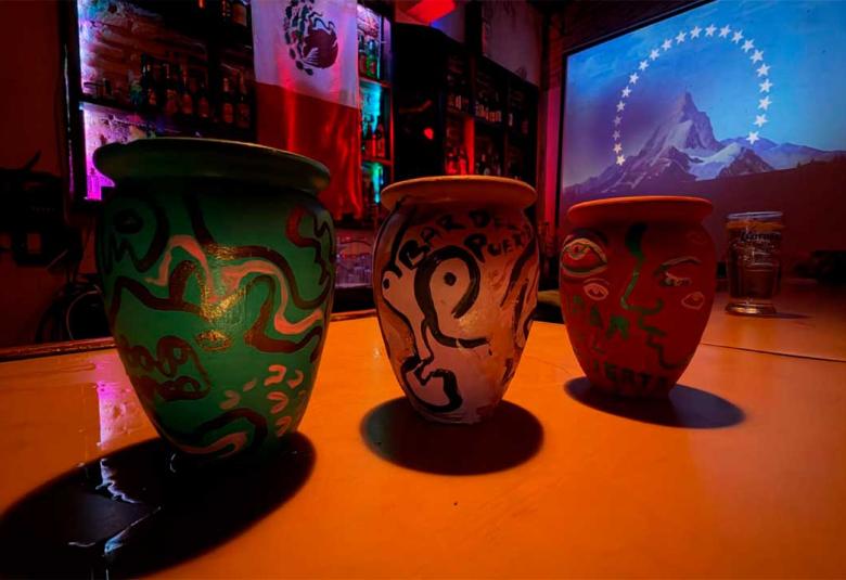 Tres cantaritos de barro pintados a mano sobre la barra del Bar del Puerto, iluminados con luces de colores y con una proyección al fondo.
