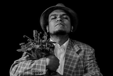 Retrato en blanco y negro del actor y director teatral Pedro Acevedo, con sombrero y saco a cuadros, sosteniendo un ramo de plantas entre sus brazos mientras mantiene los ojos cerrados en una pose introspectiva sobre un fondo negro.