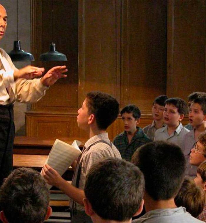 El profesor Clément Mathieu dirige un coro de niños en un salón de clases mientras varios alumnos cantan y observan atentamente durante un ensayo en una escena de la película francesa Los Coristas (Les Choristes), dirigida por Christophe Barratier.