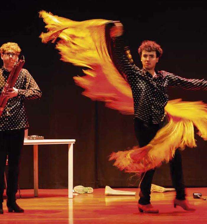 Dos artistas se presentan sobre un escenario; uno interpreta un instrumento de viento de madera mientras el otro realiza un movimiento de danza con una larga pieza de tela amarilla y naranja.