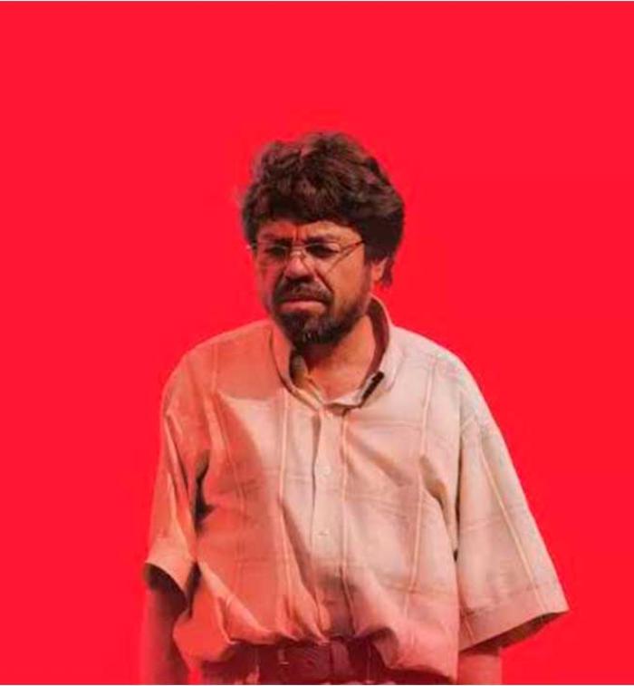 Un hombre de lentes y cabello alborotado con camisa de manga corta mirando de frente sobre un fondo rojo intenso, fotograma de la película Un Poeta de Simón Mesa Soto.